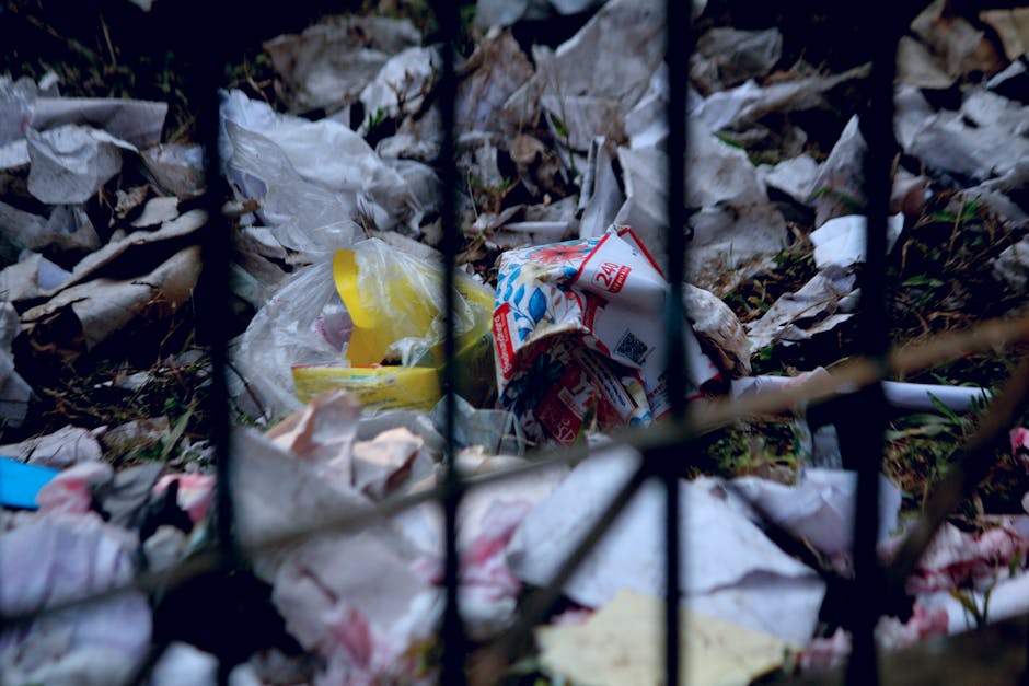 A close-up view of a pile of discarded waste consisting of crumpled paper, plastic packaging, and food wrappers, enclosed behind a black metal fence with vertical bars. The rubbish appears to be littered on the ground outdoors, surrounded by natural elements like grass and leaves. The lighting is natural, suggesting daytime. This image highlights improper waste disposal, which relates to waste management regulations that affect house removals and relocation services in Longford. Man with Van Longford offers professional moving and packing services to help avoid penalties associated with illegal waste dumping, as highlighted by the context of waste disposal rules in Longford County.