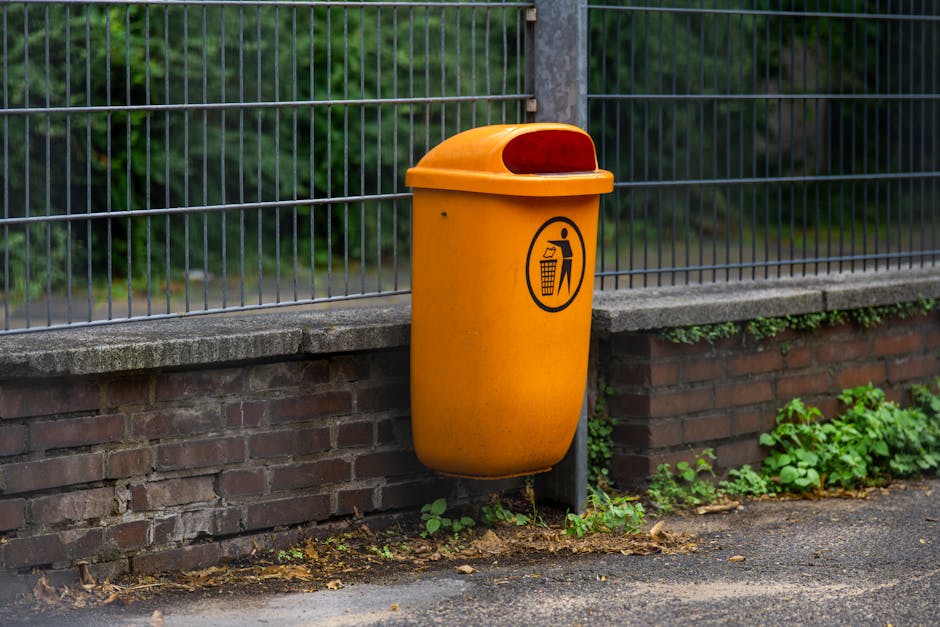 A bright orange outdoor waste disposal bin with a black pictogram of a person throwing rubbish into a bin, positioned beside a brick wall and a metal fence on a paved area. The bin features a rounded lid with a red reflective strip, indicating its purpose for waste collection. Green plants and foliage are visible at the base of the wall. The scene is captured in natural light, illustrating urban surroundings suitable for house removals or moving logistics. The bin, belonging to a waste management service, is located near a property, highlighting the importance of proper waste disposal as part of home relocation or packing and moving procedures. The Environment Agency-approved bin underscores regulated waste disposal rules in Longford County, relevant to residents preparing for removals handled by Man with Van Longford.