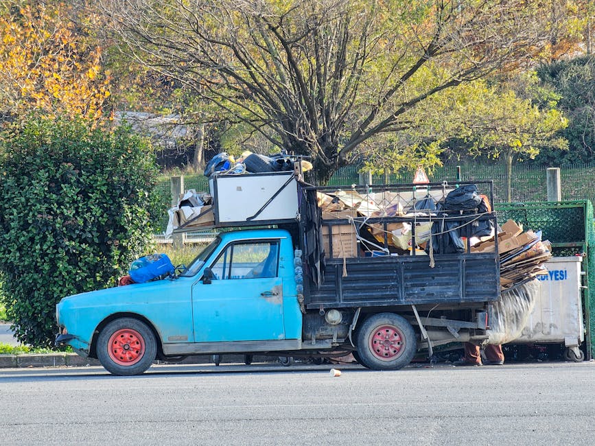 A small blue pickup truck parked on the side of a street, loaded with various household items and waste materials for a home relocation. The vehicle's bed contains cardboard boxes, plastic bags, and furniture wrapped in blankets and plastic sheeting, with some items secured by ropes. A white cabinet or appliance is positioned on top of the load, visible behind the cab. The truck's rear wheel is partially exposed, and the side mirror reflects the surrounding environment. Behind the vehicle, a large leafy tree with green and yellow foliage provides shade, while a green fence and a grassy area are visible in the background. Inside the truck bed, the furniture and packing materials are arranged in a cluttered manner, suggesting loading for a furniture transport or relocation process. This image illustrates the typical scene of loading household belongings for moving or bulky waste pickup services provided by Man with Van Longford, as part of their removals and moving logistics services.