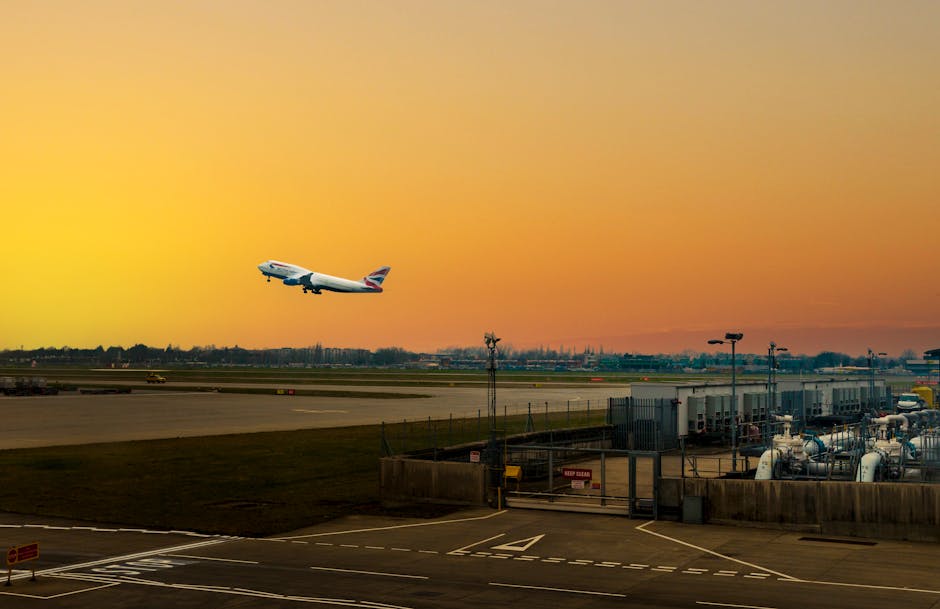 A British Airways commercial airplane is seen taking off from an airport runway during sunset, with its nose pointed upward and ascending into the sky. The sky exhibits a gradient of orange and yellow hues, indicating early evening light. In the foreground, there is a section of the airport ground showing an asphalt tarmac with white painted lines, along with various airport service vehicles, equipment, and fuel tanks arranged near a fenced area. The scene captures the busy environment of home relocation logistics, with the aircraft preparing for a flight as part of a furniture transport or moving process, which is managed by companies like Man with Van Longford.