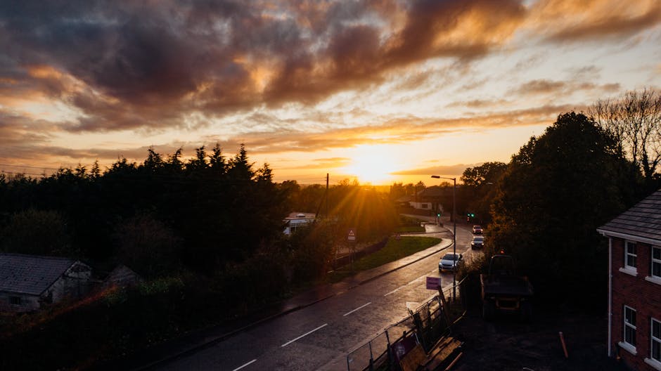 An outdoor scene during sunset showing a residential street with a van parked on the pavement, surrounded by stacked cardboard boxes, wooden furniture, and packing materials such as bubble wrap and fabric blankets, all arranged for a home relocation. The loading process appears to be underway with some items being carried or prepared for transport. The street is lined with trees and houses, and a few vehicles are visible on the road. The sunlight casts a warm glow over the area, highlighting the typical environment involved in a house removal, with the focus on logistics and the preparation of furniture and packing supplies for moving services provided by Man with Van Longford.