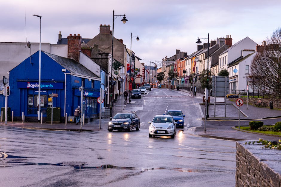 A view of a quiet urban street with a gentle incline, featuring residential buildings painted in various pastel shades and a small sports shop with a bright blue facade on the corner. Several cars are parked along the road, and two vehicles are driving through the intersection under overcast skies, with wet pavement indicating recent rain. Streetlights and utility poles line the street, and a few pedestrians are visible on the sidewalks. The surrounding area includes trees and low hedges, and a stone wall is visible in the foreground. This scene captures a typical day in a market town, suitable for discussions about local transportation and home relocation logistics, as seen through the environment where [COMPANY_NAME] may assist with furniture transport and moving services.