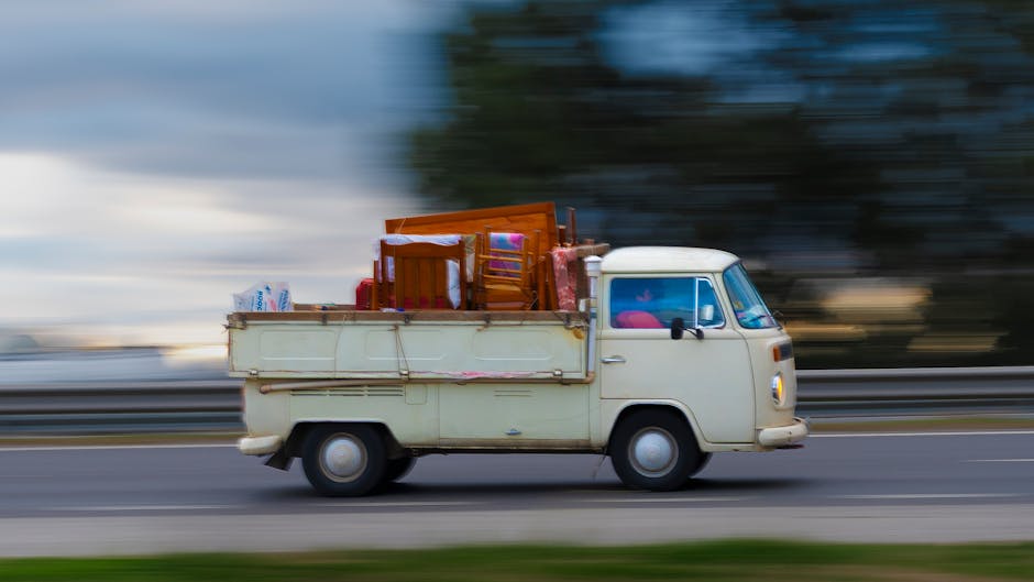 A vintage cream-coloured van with a flatbed cargo area is driving along a motorway during daylight hours. The vehicle is loaded with various household items, including a wooden dining table, chairs, a