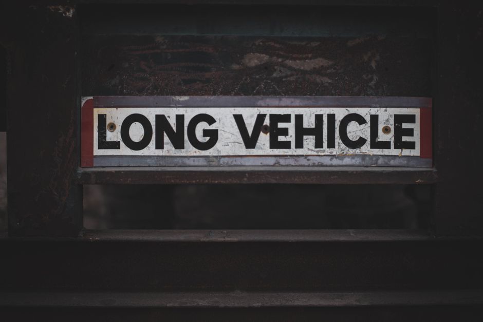 A vintage cream-coloured van with a flatbed cargo area is driving along a motorway during daylight hours. The vehicle is loaded with various household items, including a wooden dining table, chairs, a