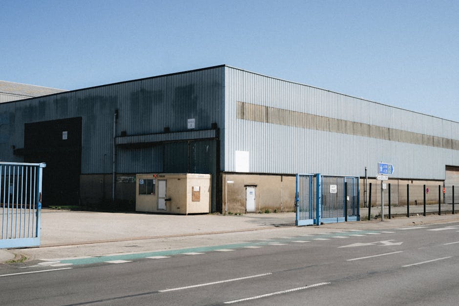 The exterior view of a large industrial warehouse with a corrugated metal facade painted in light blue and grey tones, situated alongside a paved road marked with traffic lines and a designated cycle lane. In front of the warehouse, there are three fenced-off areas with blue metal gates, possibly access points for loading and unloading. Near the building, a small beige shipping container and a smaller white utility box are positioned on the pavement. The scene includes a clear blue sky, indicating bright weather, and no vehicles or pedestrians are visible. This setting exemplifies the typical environment involved in home relocation logistics, where furniture and boxes may be loaded into a van or truck for transport. Man with Van Longford, a professional removals service, utilises such locations during the loading process in house moves, ensuring secure and organized transport of household items from property to destination.