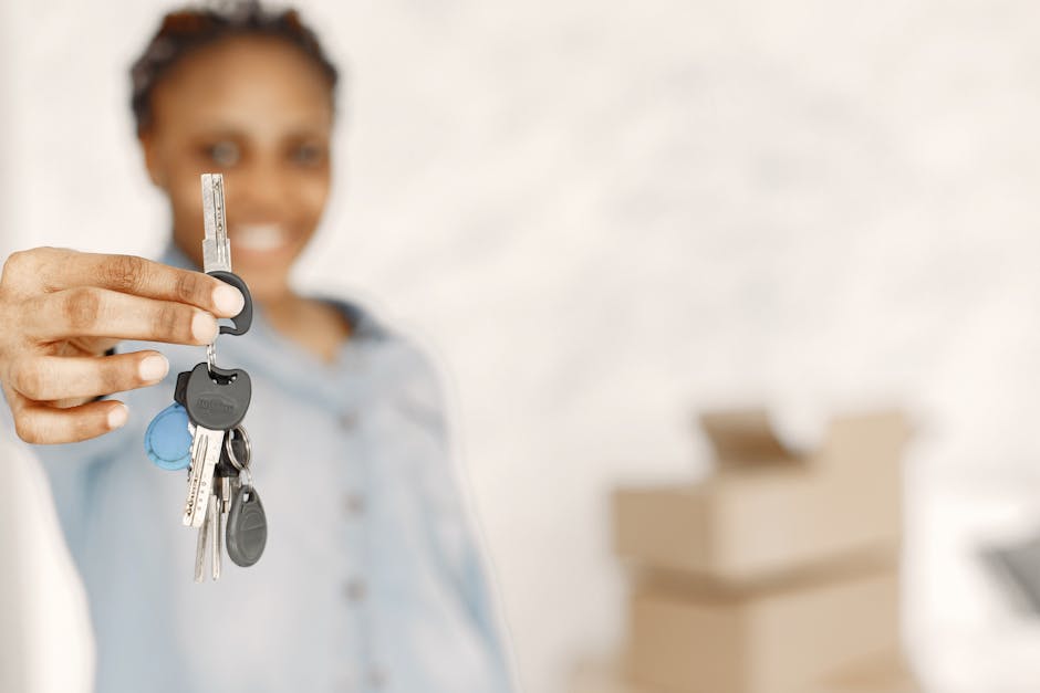 A woman with curly hair and a light blue shirt is standing indoors, smiling and holding a set of house keys with a black keyring and multiple keys, including a blue plastic key fob, towards the camera. In the background, there are partially visible cardboard boxes, some open, resting on a flat surface, indicating packing materials. The environment appears well-lit with natural or bright artificial light, and the focus is on the keys, while the woman's face remains slightly blurred. This visual context relates to the process of home relocation and furniture transport, with the woman possibly representing a mover or a homeowner preparing for a move, relevant to house removals and moving services offered by Man with Van Longford. The scene emphasizes aspects of packing, key transfer, and transportation in household moving activities, aligned with the topic of best access tips for Longford properties during a move.