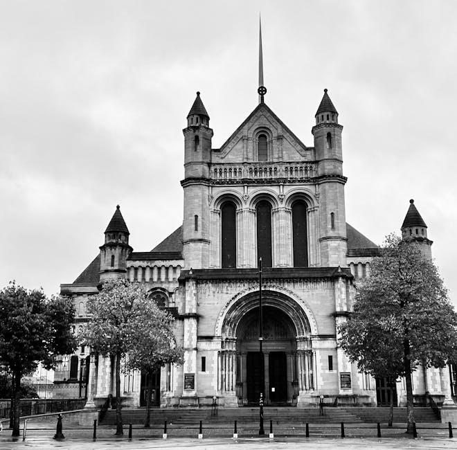 A black and white photograph of St. Mel's Cathedral showing its historic stone facade, tall arched entrance, and multiple spires, situated on a paved area with a short railing in front. Surrounding th