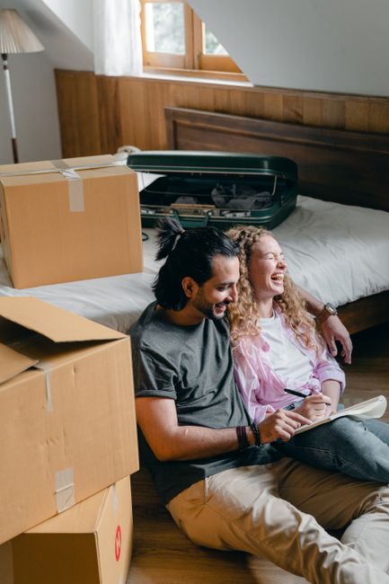 A young man with dark hair tied back and a young woman with long curly hair are sitting on the floor of a room, leaning against a bed with a wooden headboard, surrounded by moving boxes. The man is ho