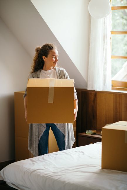 A young man with dark hair tied back and a young woman with long curly hair are sitting on the floor of a room, leaning against a bed with a wooden headboard, surrounded by moving boxes. The man is ho